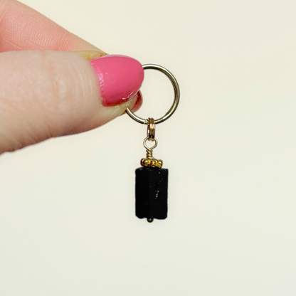 Crystal dog collar charm featuring a raw black tourmaline natural bead with gold plated accents. Featuring a brass split ring for easy attaching to your pet’s collar. Pictured being held against a cream background.