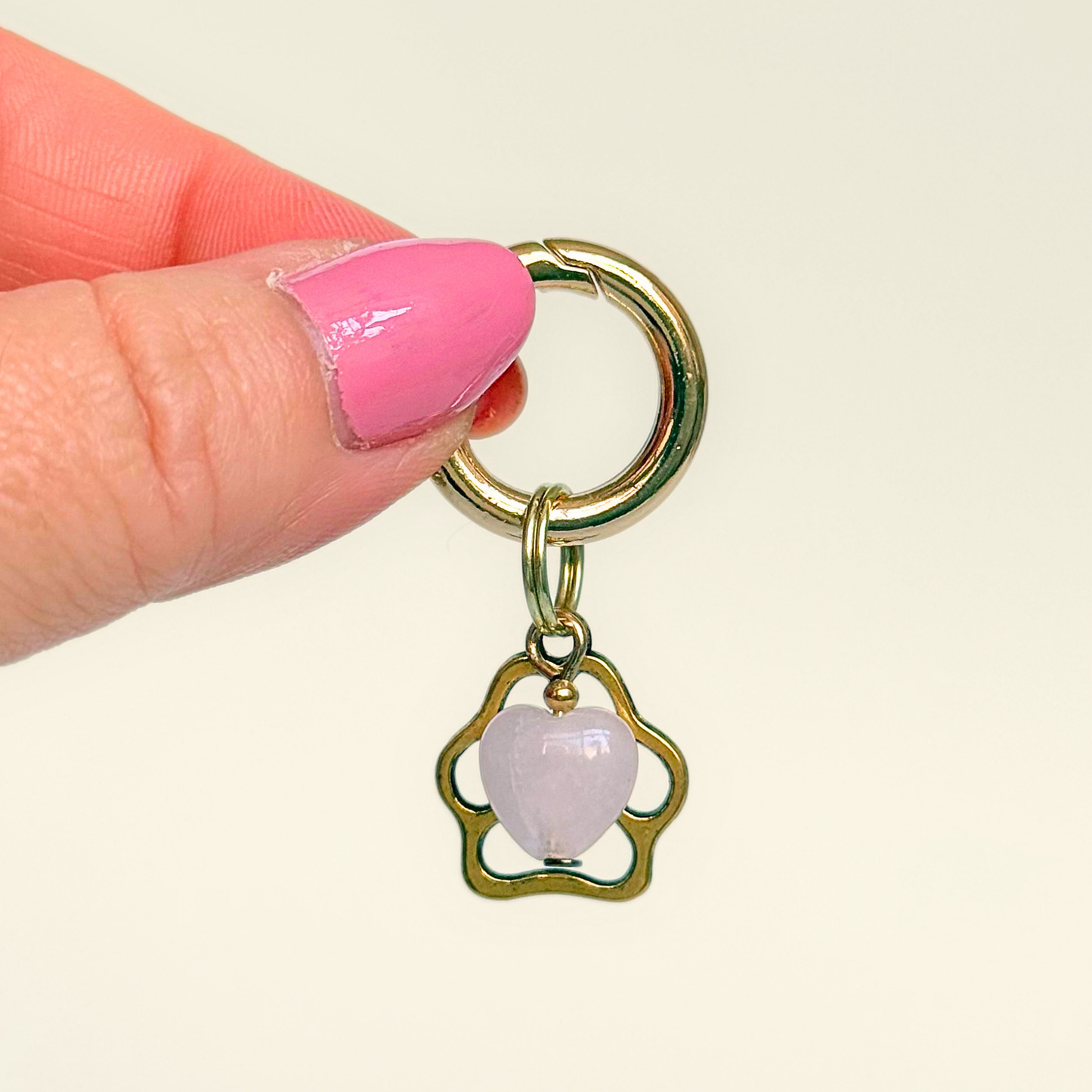 Crystal for dogs pet collar charm featuring a 10mm rose quartz heart shaped bead and a brass gold coloured paw print tag. A stone for love, calm and comfort. Pictured being held against a cream background.