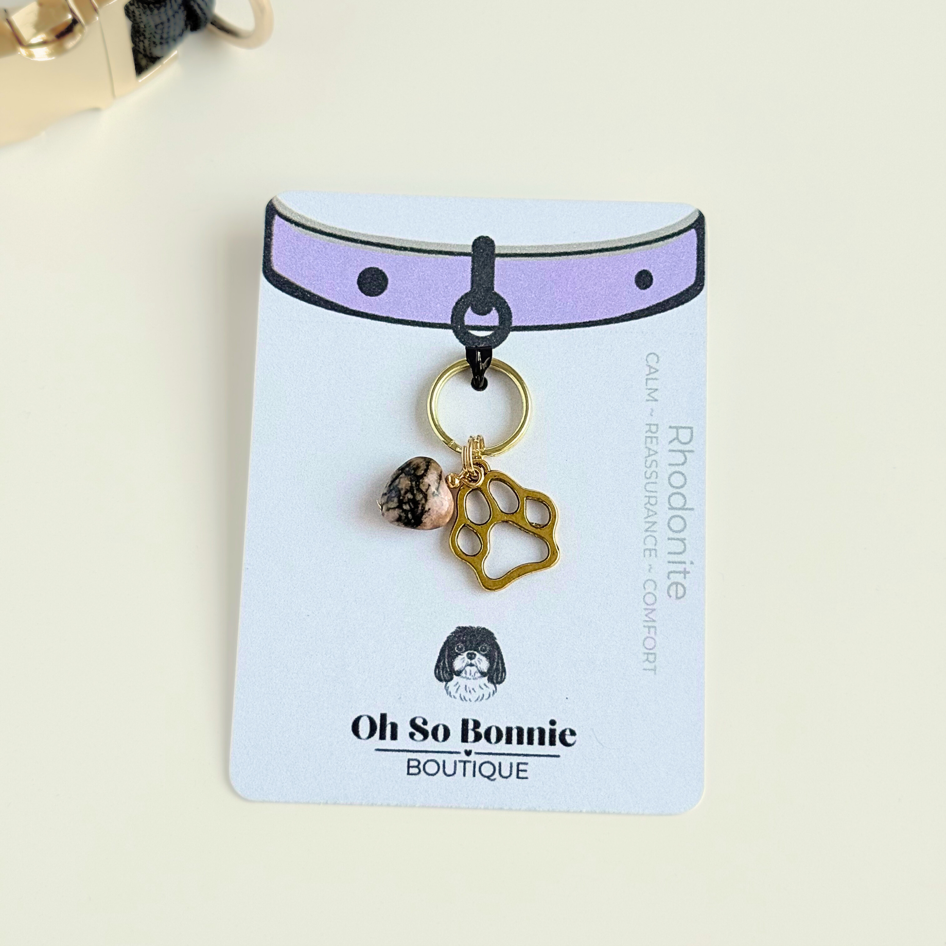 Crystal for dog collar charm featuring a 10mm rhodonite heart shaped bead and a brass gold coloured paw print tag. Pictured on the display card next to a black paracord dog collar.￼