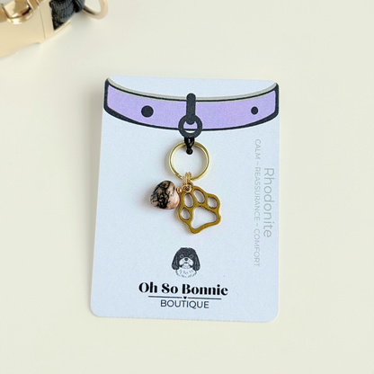 Crystal for dog collar charm featuring a 10mm rhodonite heart shaped bead and a brass gold coloured paw print tag. Pictured on the display card next to a black paracord dog collar.￼