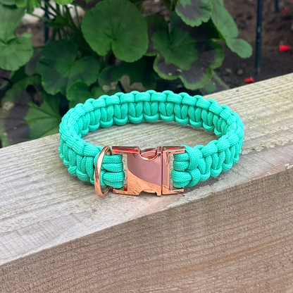 A mint green paracord braided dog collar with a rose gold metal buckle and a hanging loop, resting on a wooden deck with green garden foliage in the background.