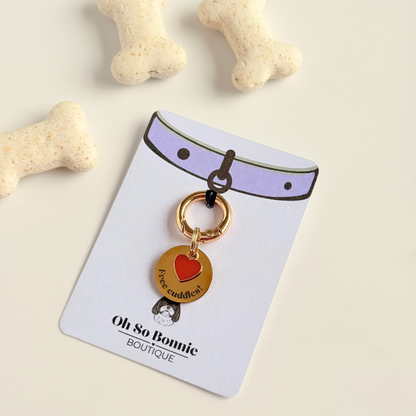 Gold plated brass and stainless steel 20mm dog collar charm with ID tag. Personalised custom identification tag with a small, red love heart charm, on a brass carabiner style o-ring. Engraved with the words ‘Free cuddles’! on the front. Pictured on the display card next to some dog bone biscuits.