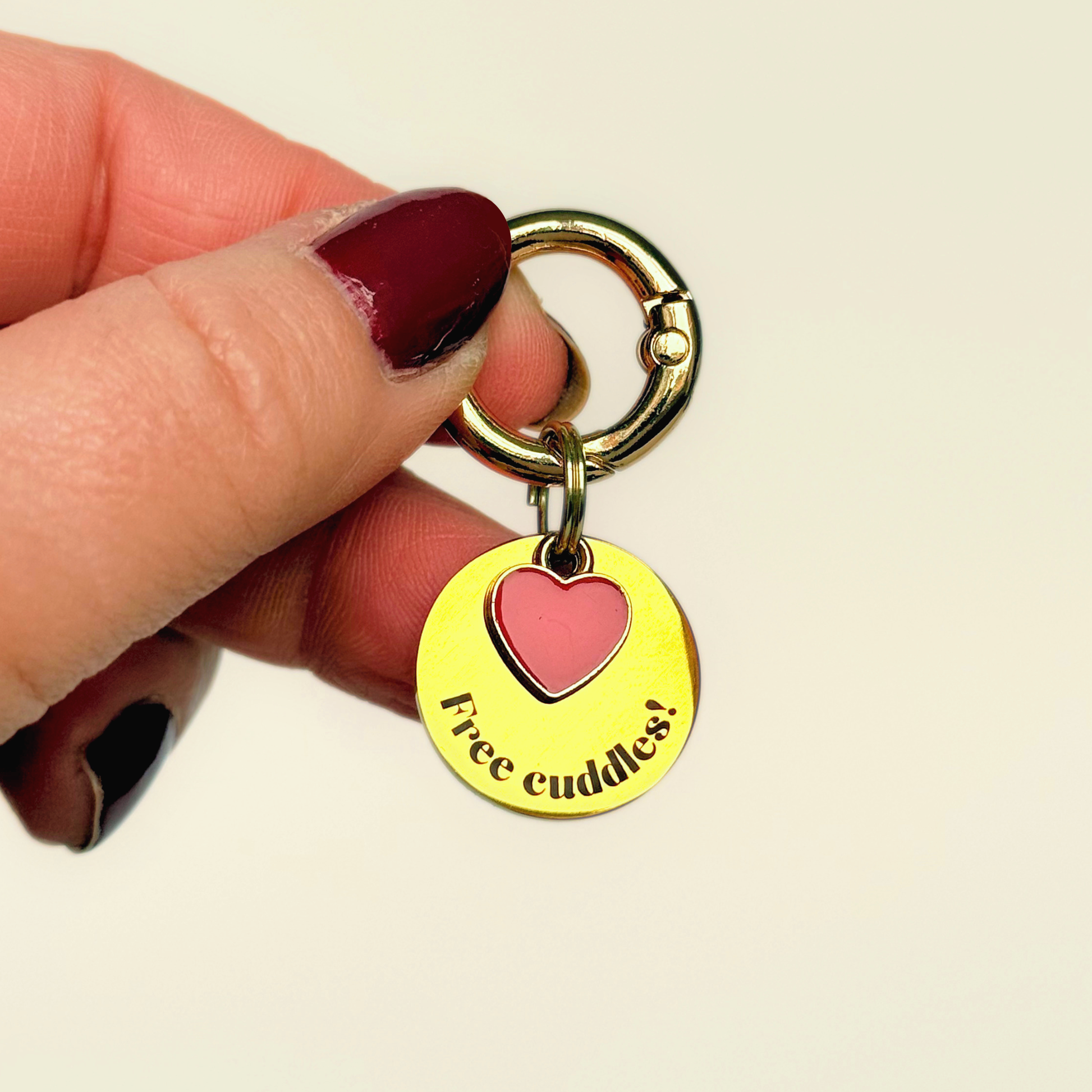 Gold plated brass and stainless steel 20mm dog collar charm with ID tag. Personalised custom identification tag with a small, red love heart charm, on a  brass carabiner style o-ring. Engraved with the words ‘Free cuddles’! on the front. Pictured being held up against a cream background.