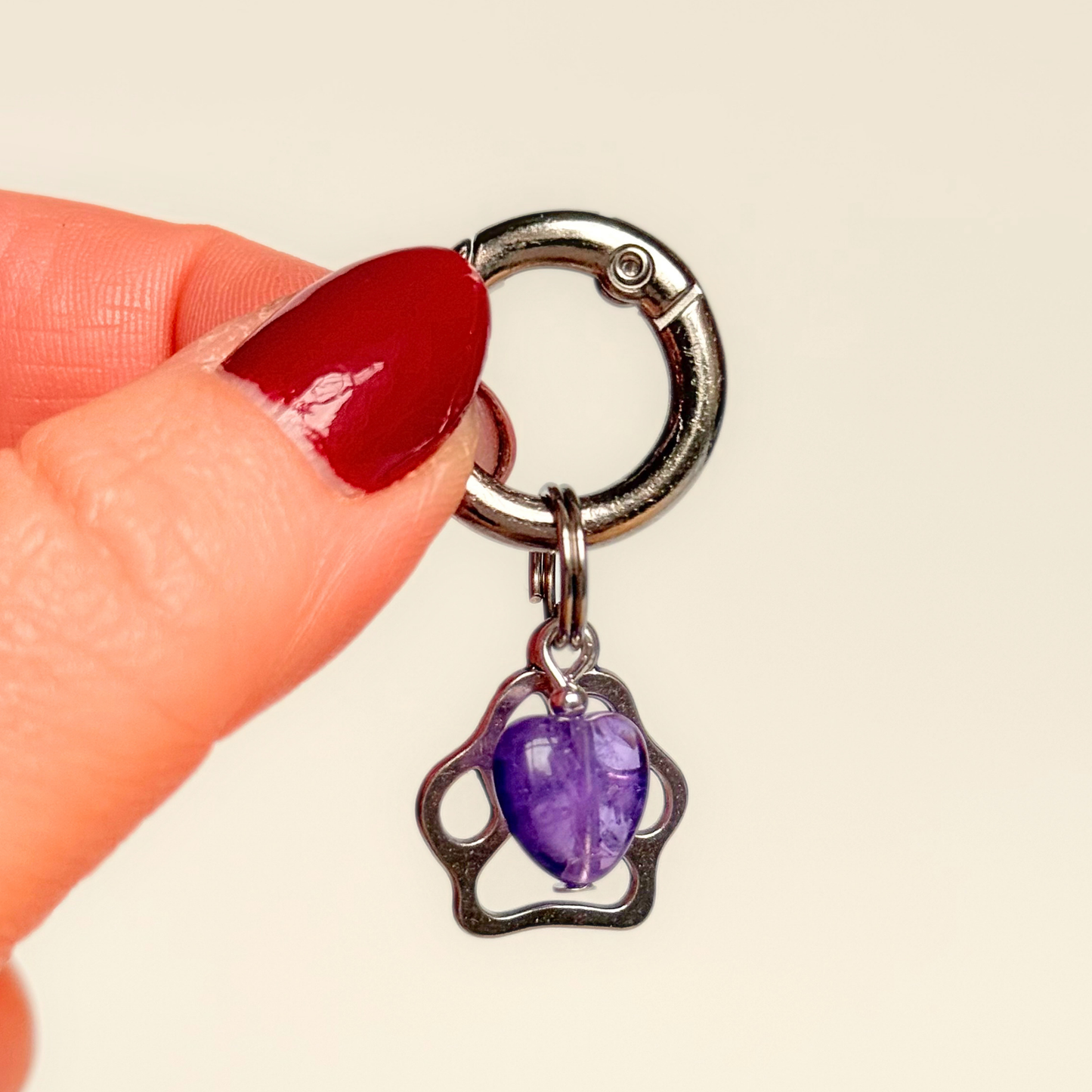 Crystal for dogs pet collar charm featuring a 10mm purple amethyst heart shaped bead and a brass silver coloured paw print tag. Pictured being held against a cream background.