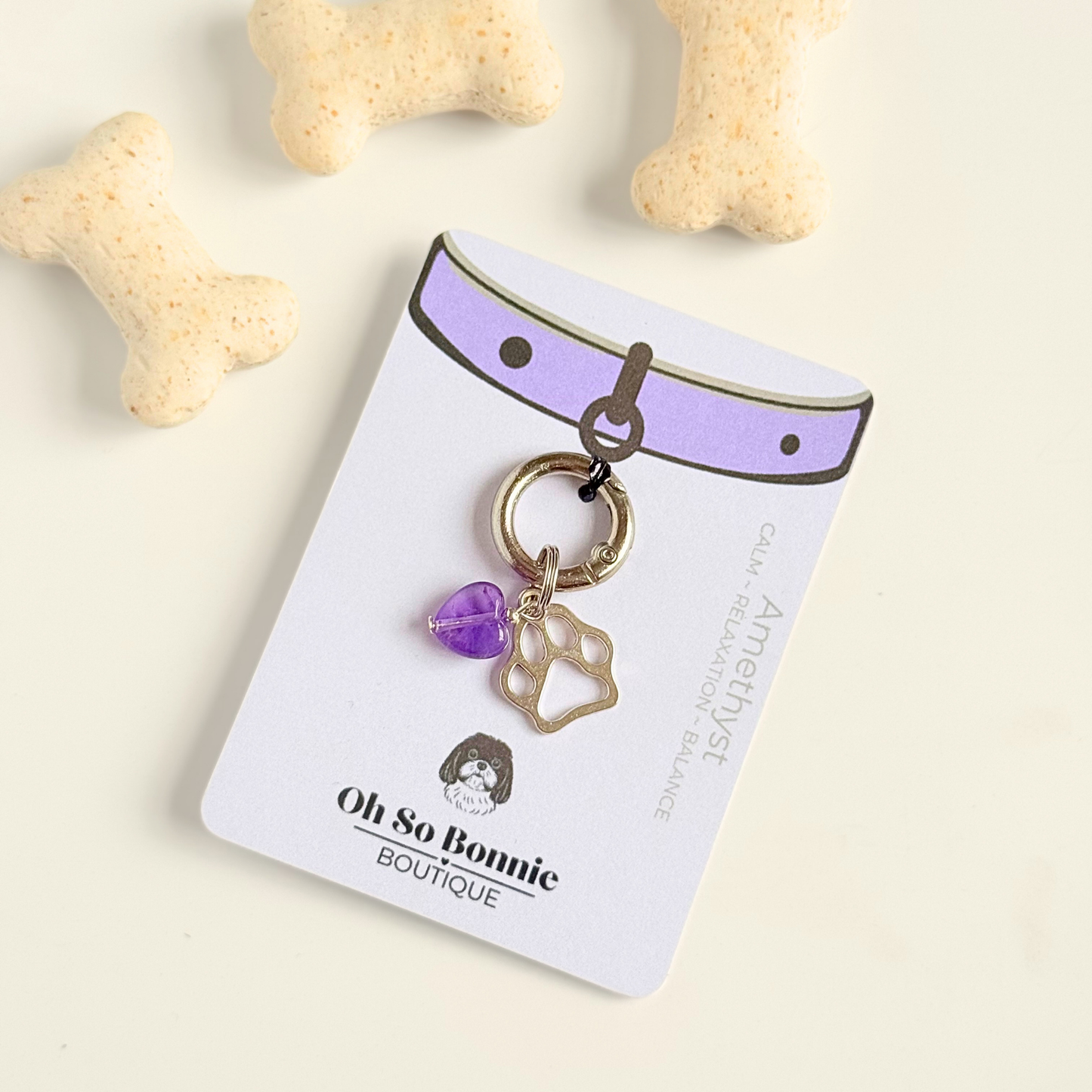 Crystal for dogs pet collar charm featuring a 10mm purple amethyst heart shaped bead and a brass silver coloured paw print tag. Pictured on the display card, next to some dog biscuits.