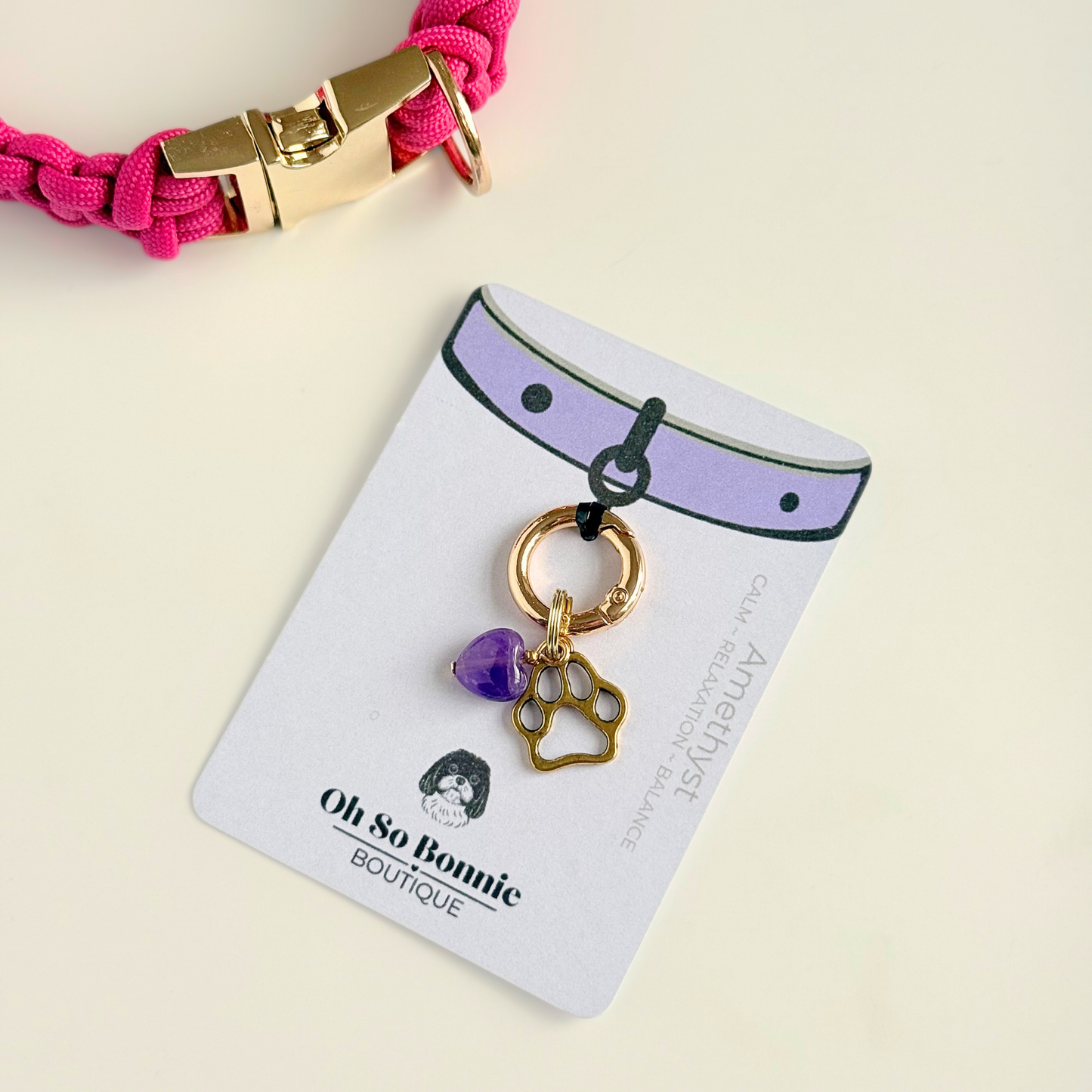 Crystal for dogs pet collar charm featuring a 10mm purple amethyst heart shaped bead and a brass gold coloured paw print tag. Pictured on the isplay card, next to a pink paracord dog collar.
