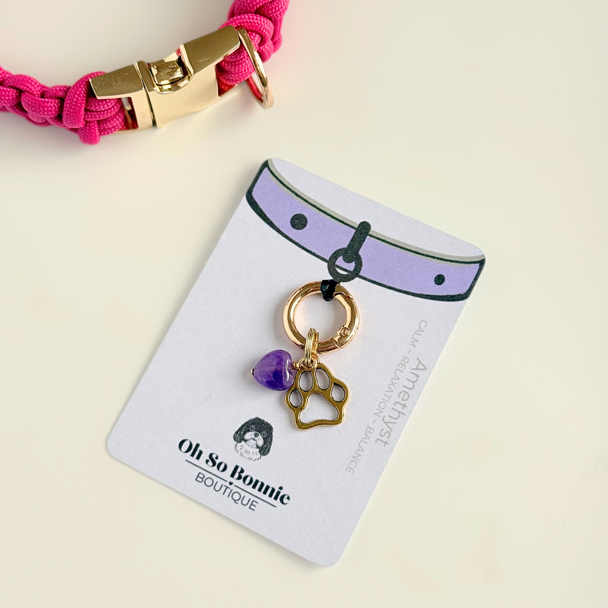 Crystal for dogs pet collar charm featuring a 10mm purple amethyst heart shaped bead and a brass gold coloured paw print tag. Pictured on the isplay card, next to a pink paracord dog collar.