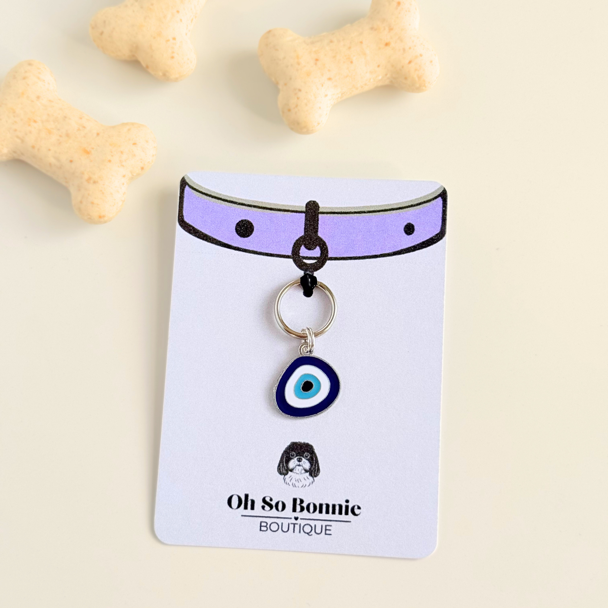 Silver plated blue turkish evil eye dog collar charm, for protection, security and grounding, 35mm easy to attach to pet collars. Picture shows the tag on the display card, next to some dog biscuits.