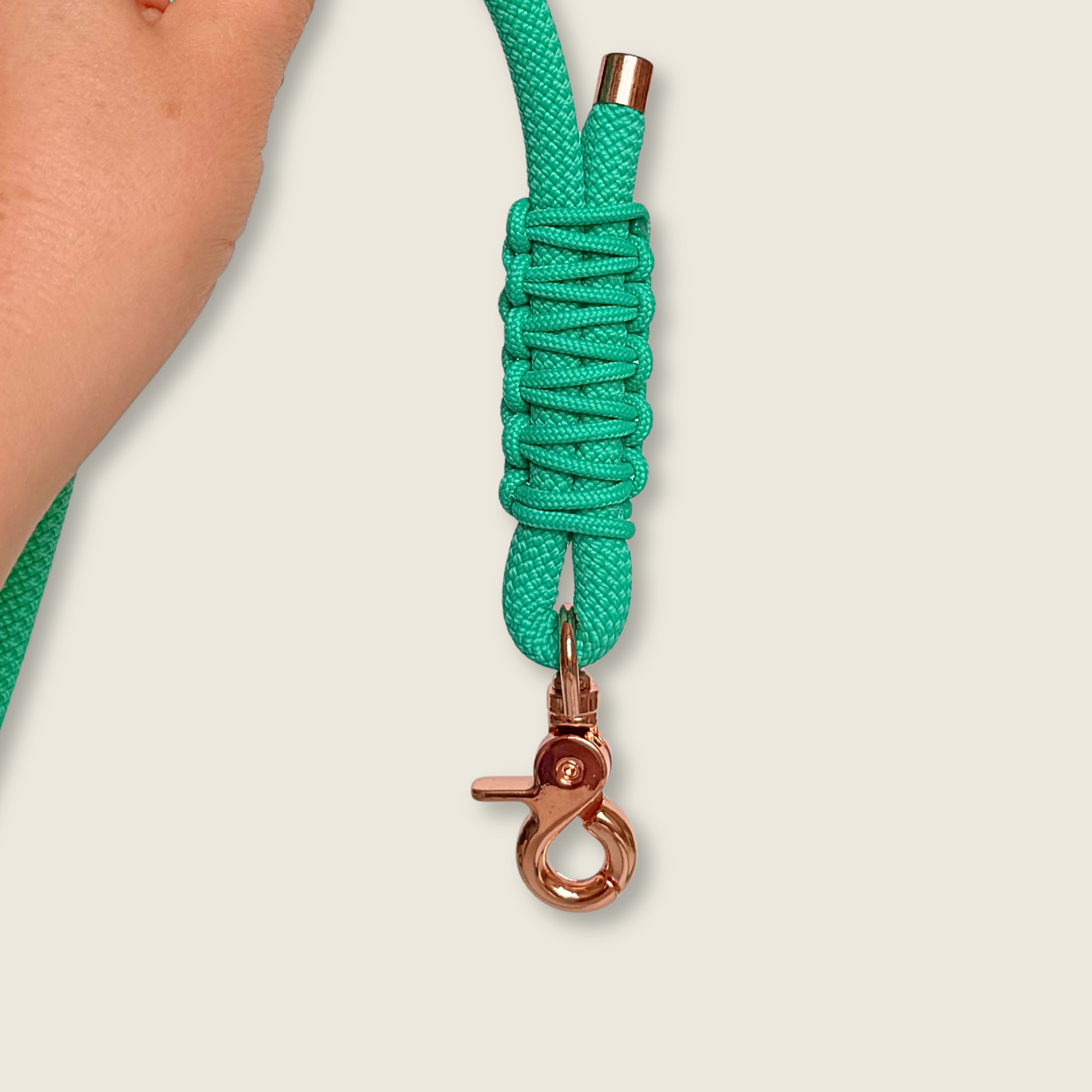 A close-up of a bright mint green braided rope dog leash wrapped around a hand and attached to a rose gold metal swivel clasp, against a light beige background.