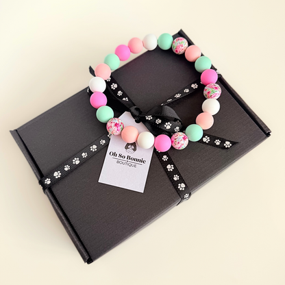 Silicone beaded dog necklace made with 15mm round beads in pink, green, white and a floral flowers print. On a strong, elastic stretch cord. Pictured on top of the gift box.￼