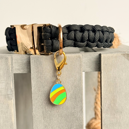 Colourful striped Easter egg enamel dog collar charm that easily attaches to collars and harnesses with a gold plated lobster clasp. Close-up picture of the charm attached to a black paracord dog collar.￼