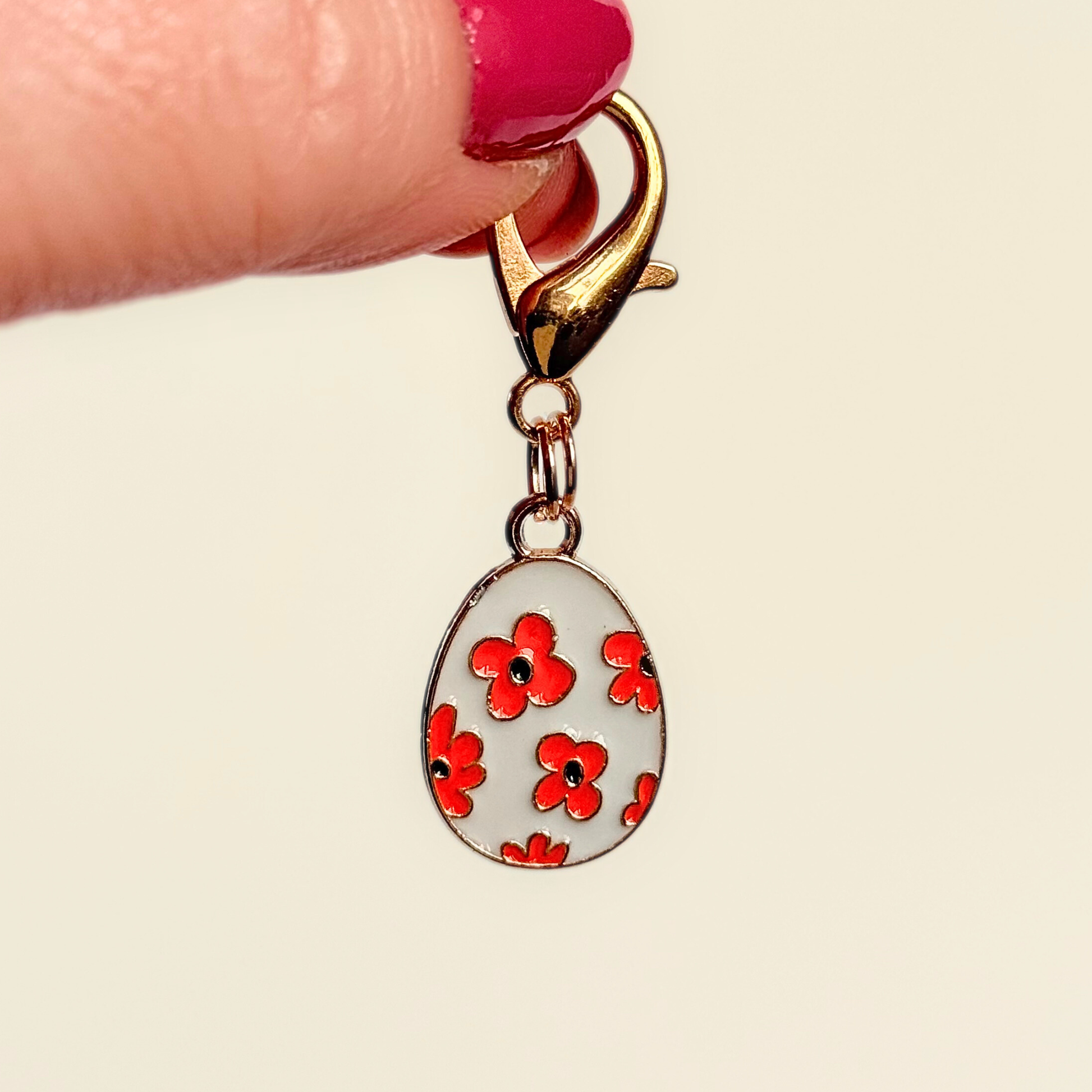 White with red flowers Easter egg enamel dog collar charm that easily attaches to collars and harnesses with a gold plated lobster clasp. Pictured being held up against a cream background.￼