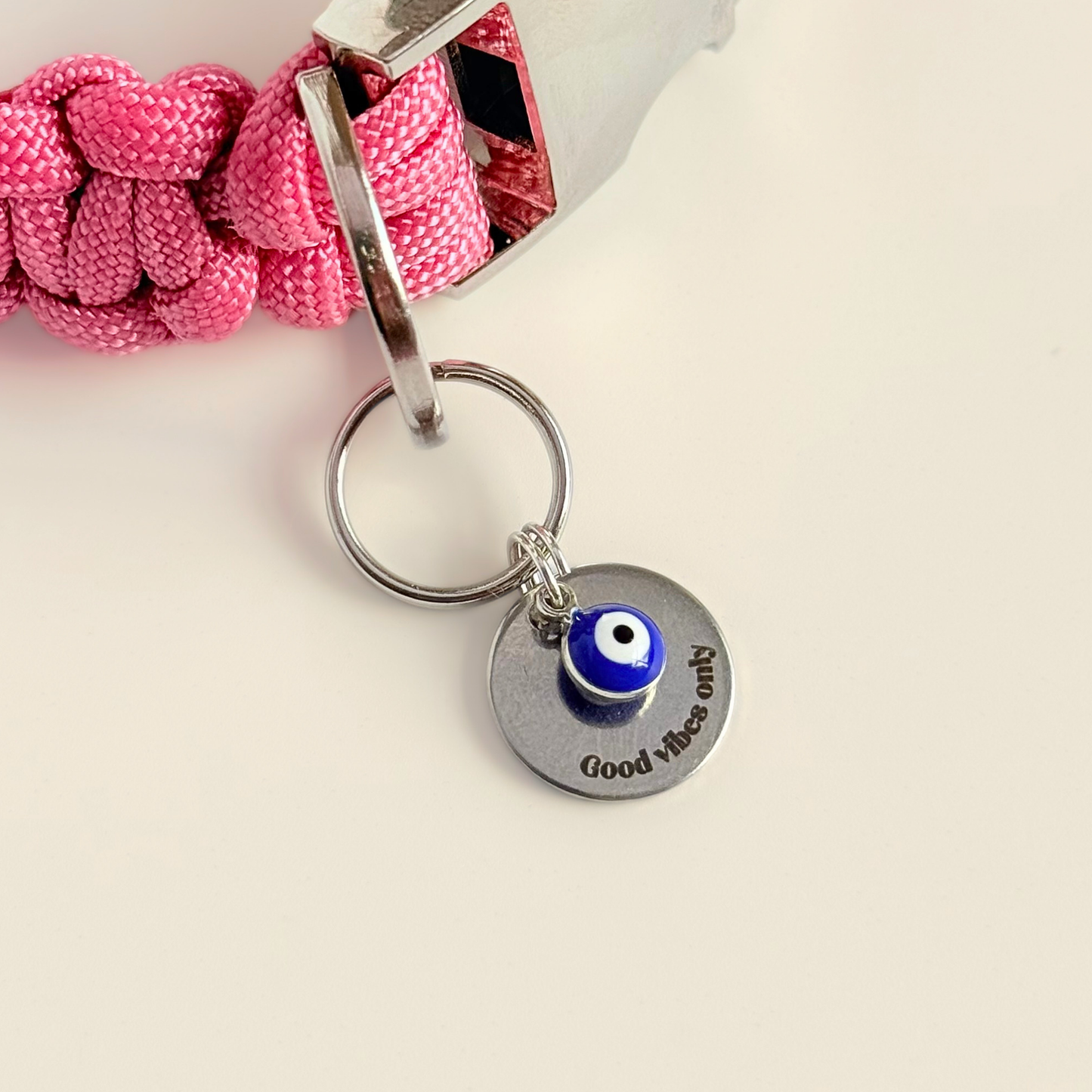 Silver plated brass and stainless steel 15mm dog collar charm with ID tag. Personalised custom identification tag with a small, blue Turkish Evil Eye charm, on a 15mm silver split ring. Engraved with the words ‘Good vibes only’ on the front. Pictured attached to a pink collar with a cream background.