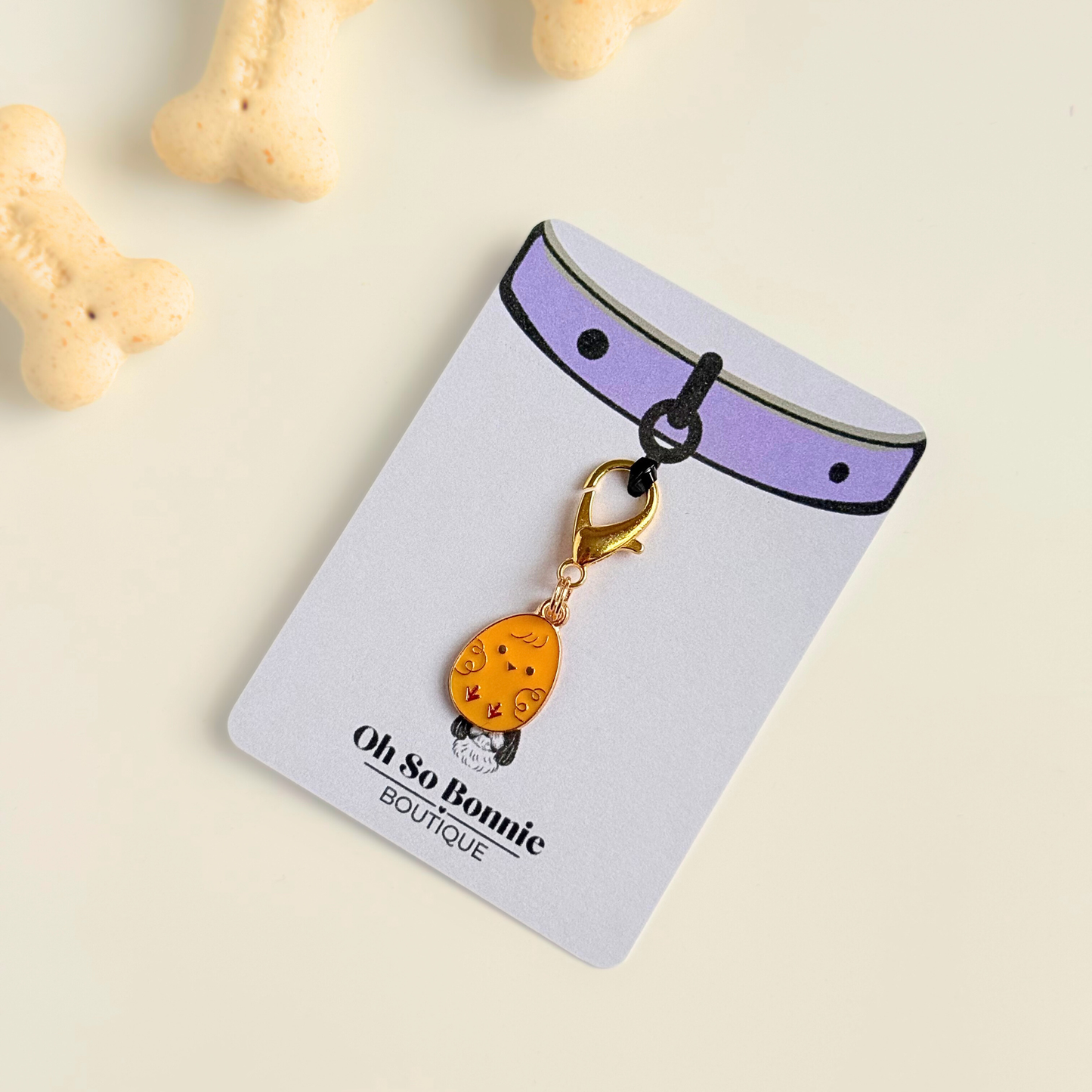 Yellow chick Easter egg enamel dog collar charm that easily attaches to collars and harnesses with a gold plated lobster clasp. Pictured on the display card, next to dog biscuits.