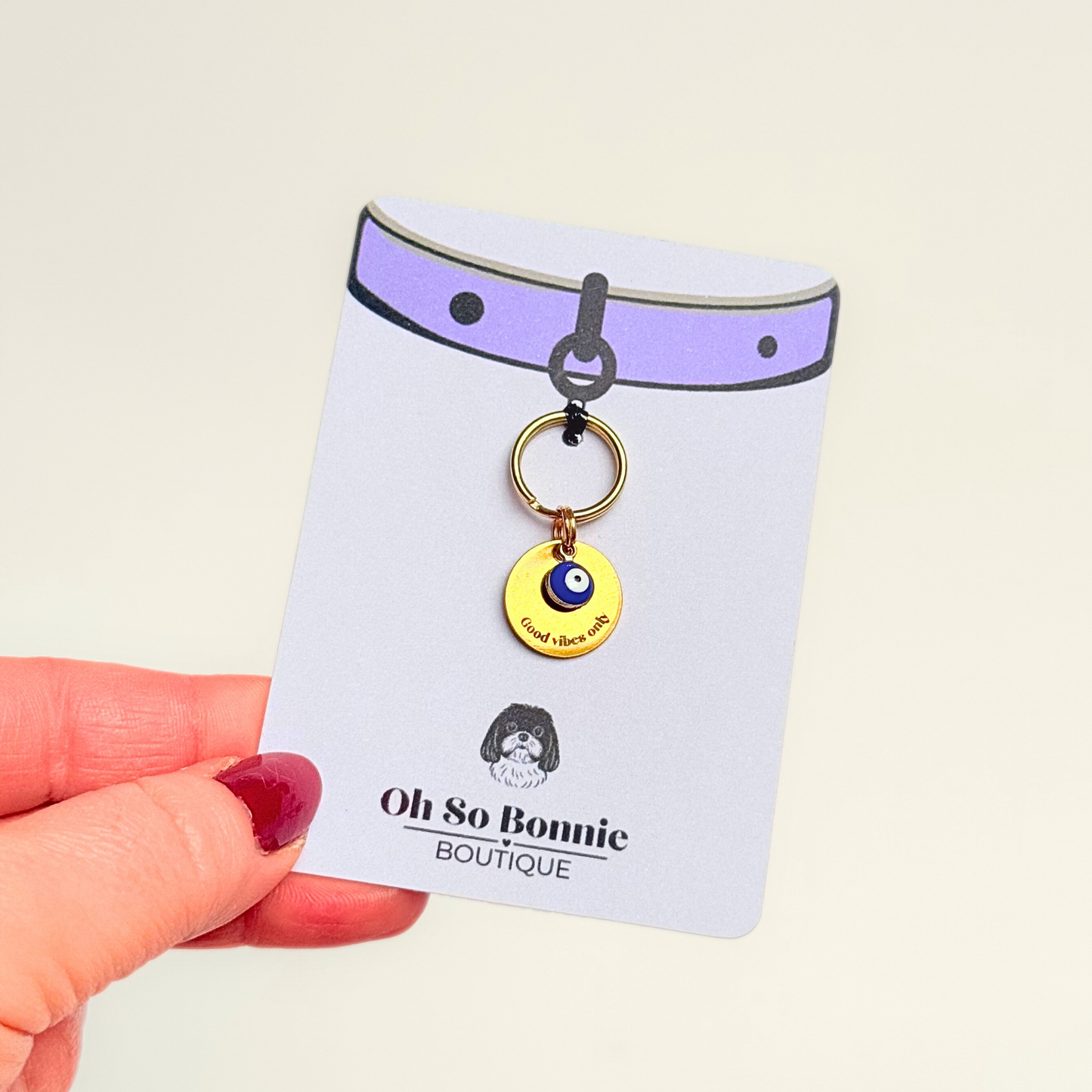 Gold plated brass and stainless steel 15mm dog collar charm with ID tag. Personalised custom identification tag with a small, blue Turkish Evil Eye charm, on a 15mm brass split ring. Engraved with the words ‘Good vibes only’ on the front. Pictured on the display card being held up.