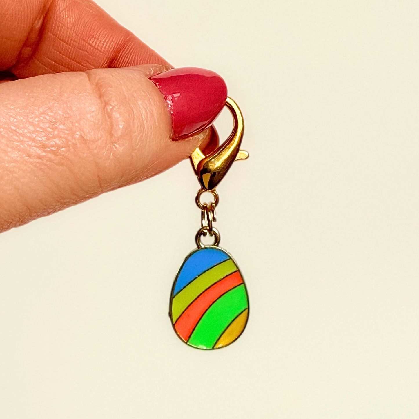 Colourful striped Easter egg enamel dog collar charm that easily attaches to collars and harnesses with a gold plated lobster clasp. Pictured being held against a cream background.