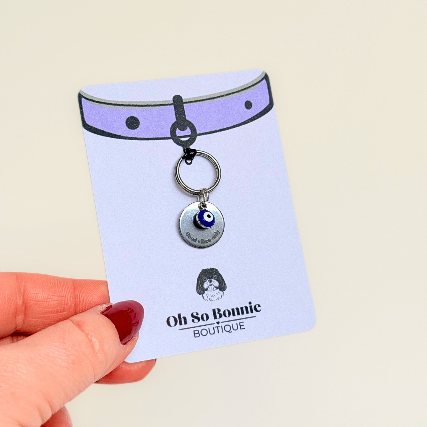 Silver plated brass and stainless steel 15mm dog collar charm with ID tag. Personalised custom identification tag with a small, blue Turkish Evil Eye charm, on a 15mm silver split ring. Engraved with the words ‘Good vibes only’ on the front. Pictured on the display card being held up.