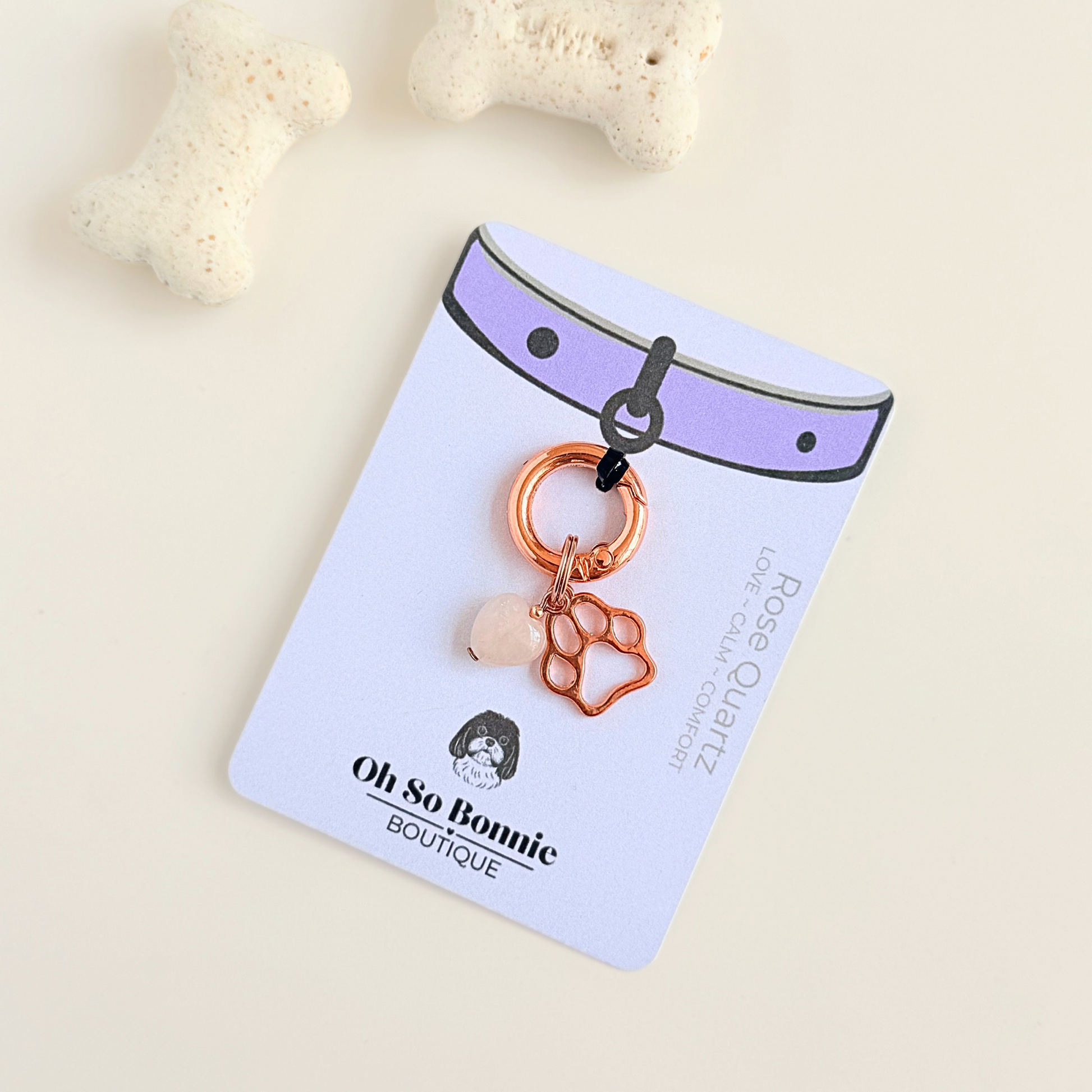 Crystal for dogs pet collar charm featuring a 10mm rose quartz heart shaped bead and a brass rose-gold coloured paw print tag. A stone for love, calm and comfort. Pictured on the display card, next to some dog bone biscuits