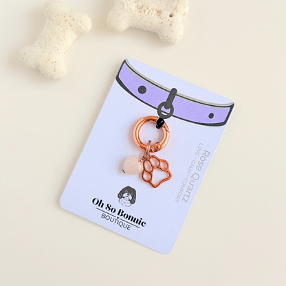 Crystal for dogs pet collar charm featuring a 10mm rose quartz heart shaped bead and a brass rose-gold coloured paw print tag. A stone for love, calm and comfort. Pictured on the display card, next to some dog bone biscuits