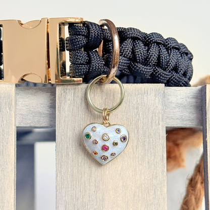 Heart shaped cream / off white pearlescent coloured enamel and brass pet collar charm featuring a selection of multicoloured cubic zirconia stones.  Pictured attached to a black paracord collar, close-up.