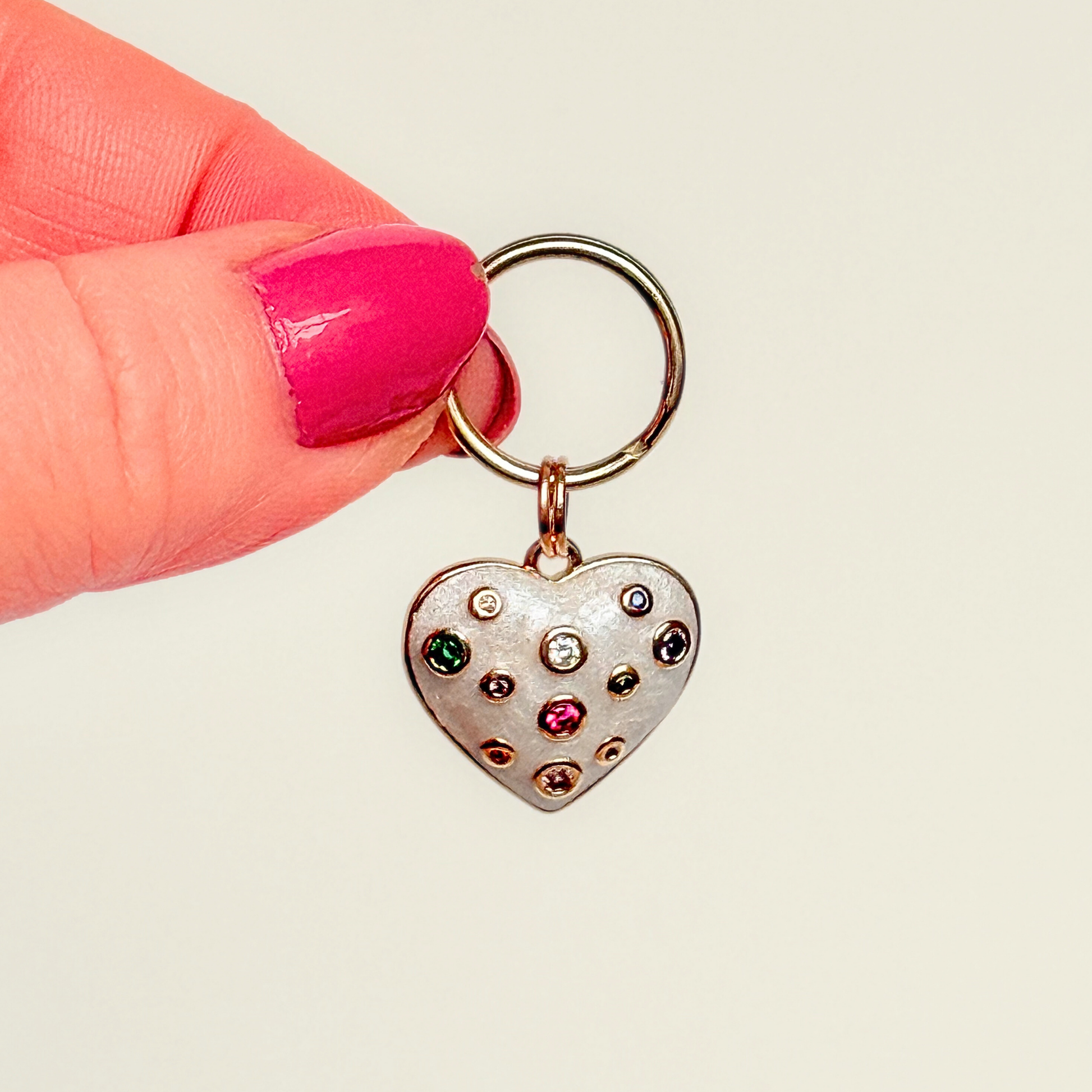 Heart shaped cream / off white pearlescent coloured enamel and brass pet collar charm featuring a selection of multicoloured cubic zirconia stones. Pictured being held, against a cream background.