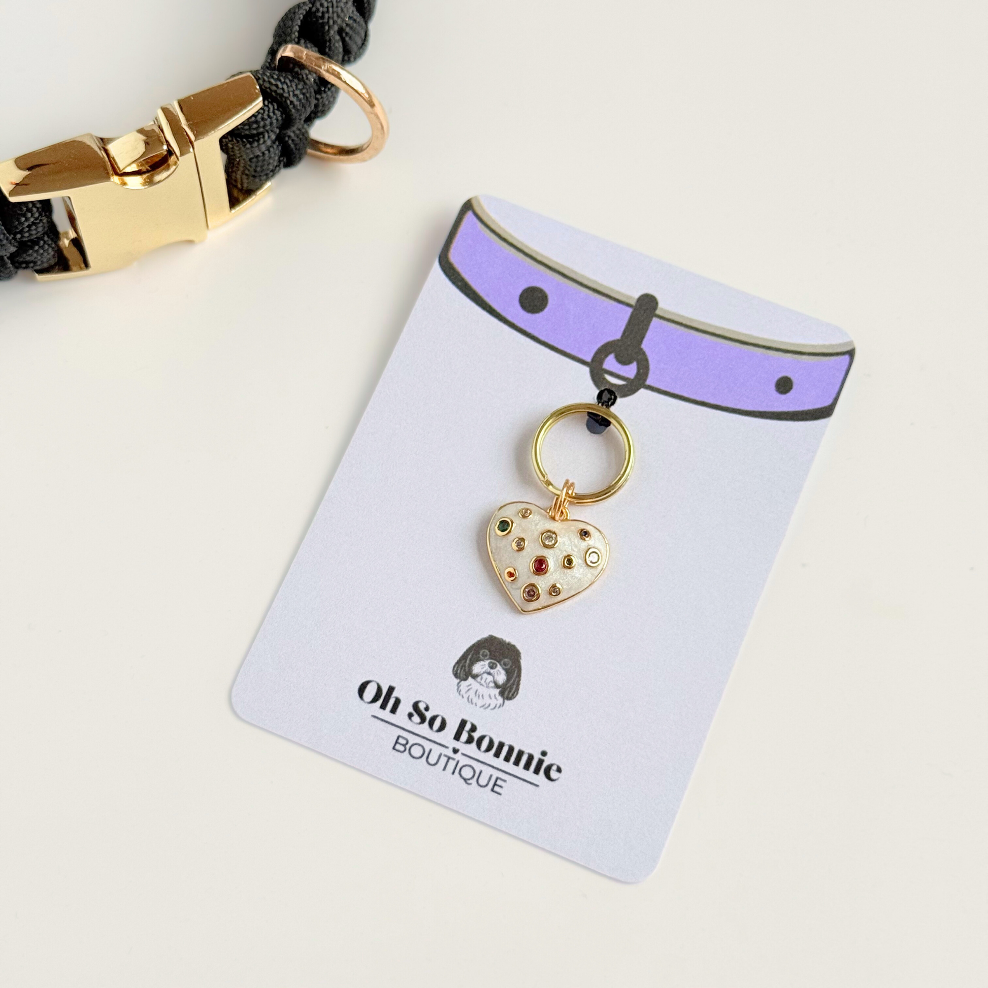 Heart shaped cream / off white pearlescent coloured enamel and brass pet collar charm featuring a selection of multicoloured cubic zirconia stones. Pictured on the display card next to a black collar.