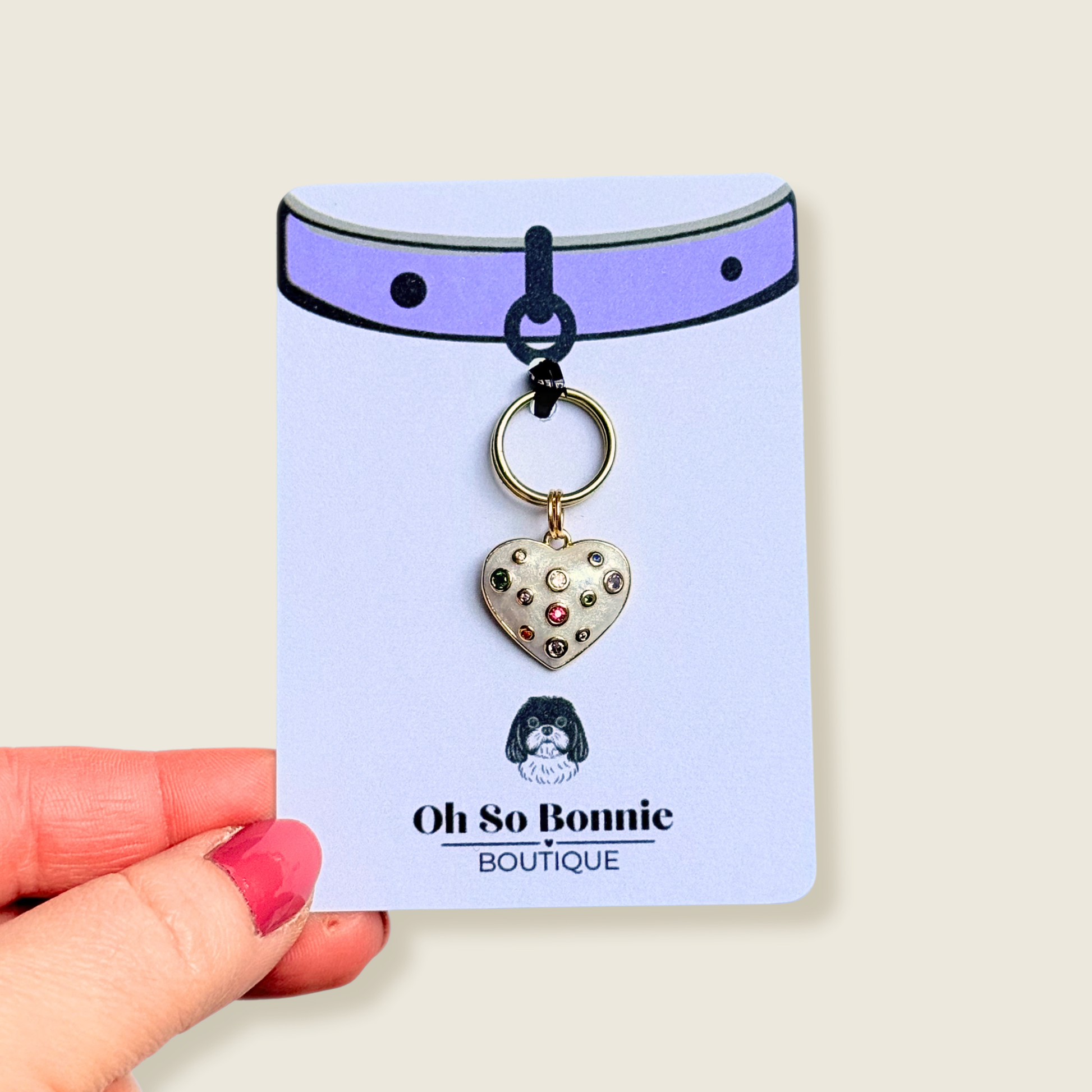 Heart shaped cream / off white pearlescent coloured enamel and brass pet collar charm featuring a selection of multicoloured cubic zirconia stones. Pictures on the display card, being held against a cream background.