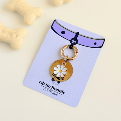 Gold white daisy flower dog collar ID tag, Enamel floral charm with dog name and address details. easily clips onto collar or harness. Pictured on the display card, next to some dog biscuits.