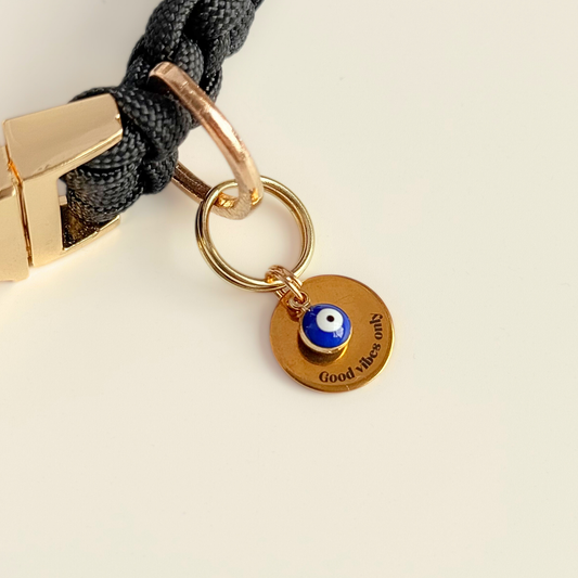 Gold plated brass and stainless steel 15mm dog collar charm with ID tag. Personalised custom identification tag with a small, blue Turkish Evil Eye charm, on a 15mm brass split ring. Engraved with the words ‘Good vibes only’ on the front. Pictured attached to a black collar with a cream background.