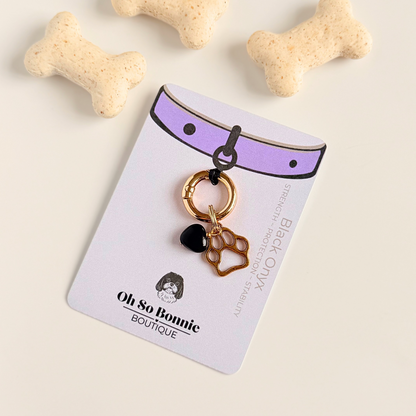 Crystal for dogs pet collar charm featuring a 10mm black onyx heart shaped bead and a brass gold coloured paw print tag. A stone for protection, strength and stability. Pictured on the display card, next to some dog biscuits 