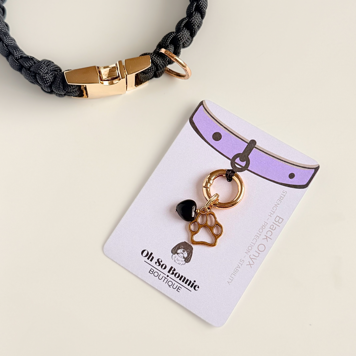 Crystal for dogs pet collar charm featuring a 10mm black onyx heart shaped bead and a brass gold coloured paw print tag. A stone for protection, strength and stability. Pictured on the display card, next to a black paracord dog collar