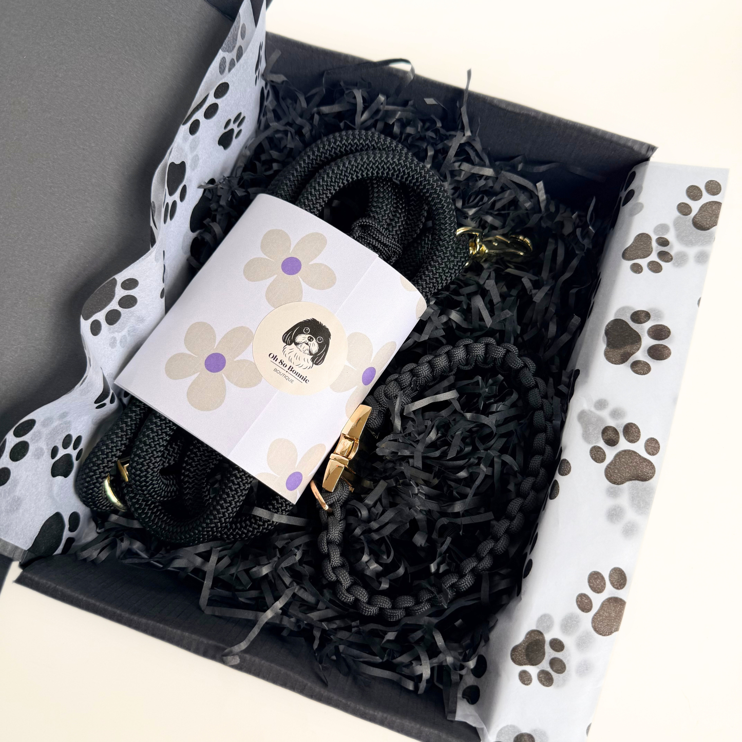 A black dog lead and collar set presented in a black gift box. The box is filled with black shredded paper and paw print tissue. A white label with a floral design and a dog illustration is wrapped around the lead.