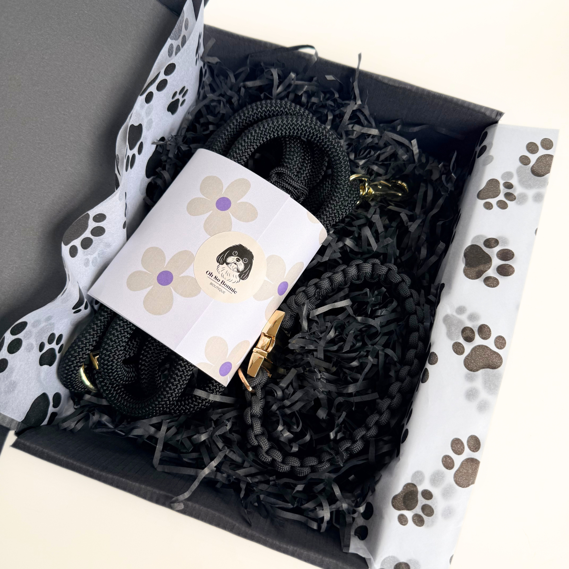 A black dog lead and collar set presented in a black gift box. The box is filled with black shredded paper and paw print tissue. A white label with a floral design and a dog illustration is wrapped around the lead.